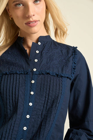 A close-up of a woman wearing a navy blue long-sleeve blouse with a high ruffled collar, vertical pleated front panel, and white buttons, against a light beige background.