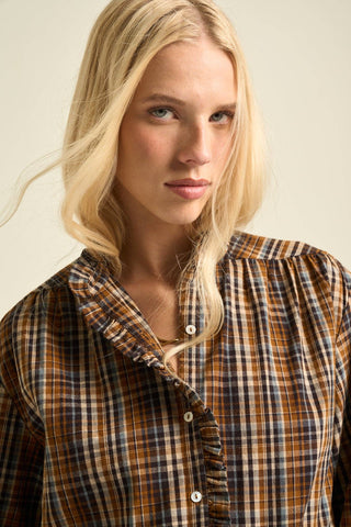 A close-up shot of a blonde woman wearing a brown, beige, and navy plaid button-up shirt with ruffled details on the collar and cuffs, set against a plain beige background.