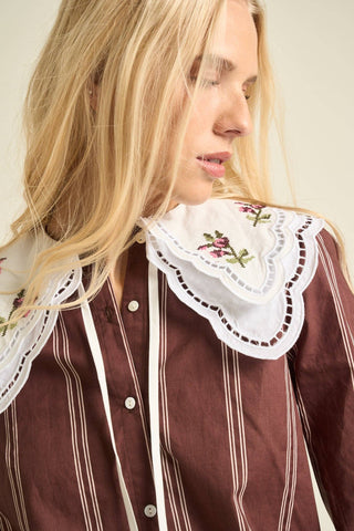 A close-up of a woman wearing a brown and white vertically striped shirt with a large white embroidered lace collar.