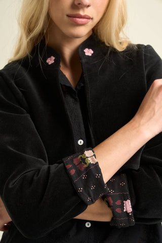 A close-up of a model wearing a black velvet button-up shirt with pink floral embroidery on the collar and a matching patterned fabric cuff.