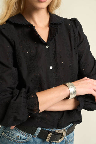 A close-up of a woman wearing a black long-sleeve button-up blouse with eyelet embroidery, ruffled collar and cuffs, tucked into blue jeans with a black belt.