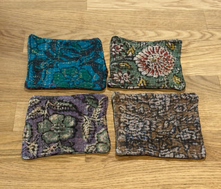 Four small square fabric pouches or coasters with different batik patterns, arranged in a 2x2 grid on a light oak wooden tabletop.