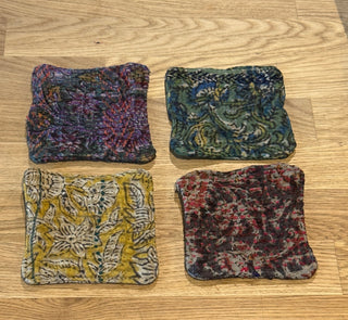 Four square fabric coasters with colorful floral and abstract patterns, arranged in a 2x2 grid on a light wood countertop above white cabinets.