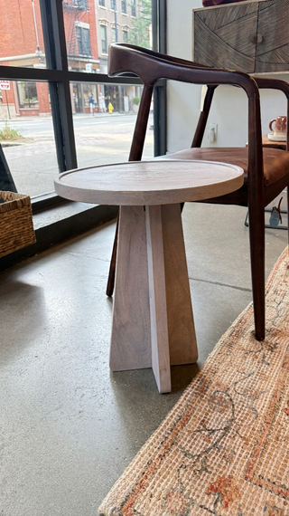 A light natural wood side table with a round top and tapered pedestal base, styled in a modern loft setting next to a dark wood chair.