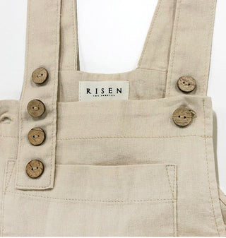 Close-up of a beige linen bib overalls with brown wooden buttons and a white brand label reading 'RISEN LOS ANGELES' on a white background.
