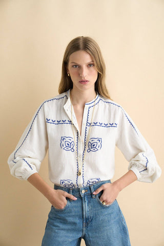 Woman wearing a white blouse with blue embroidery and blue jeans against a beige background