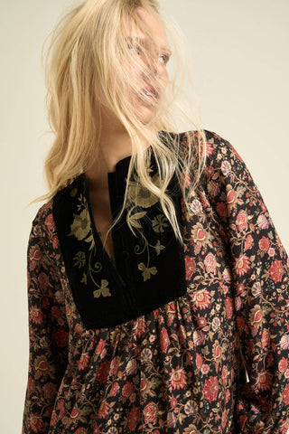 A close-up of a woman wearing a long-sleeve bohemian-style dress with a black velvet embroidered yoke and a red and gold floral print on a black background.