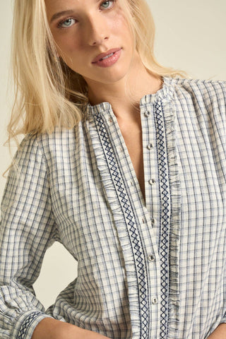 A close-up of a woman wearing a long-sleeve button-front blouse with a white, gray, and navy plaid pattern and decorative embroidered trim.