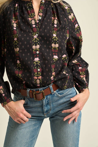 A close-up of a person wearing a black long-sleeve blouse with a colorful floral and geometric vertical stripe pattern, tucked into blue jeans with a brown belt.
